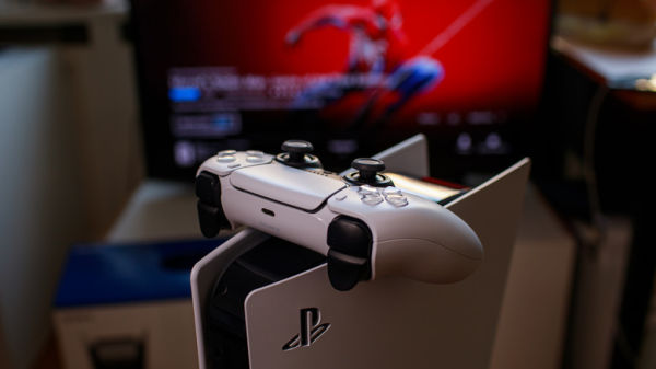 A PlayStation 5 sitting upright with a DualSense controller on top of it and SpiderMan on the televsion screen in the background