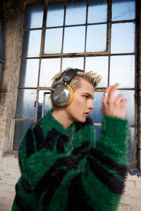 This may contain: a young man with headphones standing in front of a window wearing a green sweater