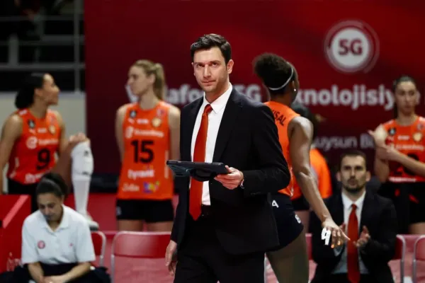 Turkish volleyball coach Ferhat Akbas standing courtside in a black suit with an orange tie, holding a tablet and appearing focused.