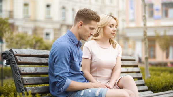 A young couple sits silently on a park bench, appearing upset and awkward.