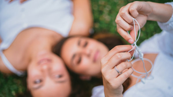 Two people lying in the grass, one holding white wired Apple headphones up to the camera to untangle them.