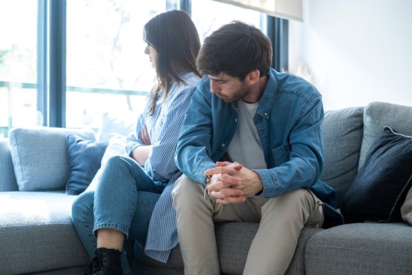 A couple sits on a couch, turned away from each other, appearing upset.
