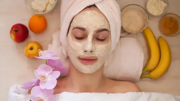 : A woman is lying down with her eyes closed, wearing a creamy face mask and a towel wrapped around her head.