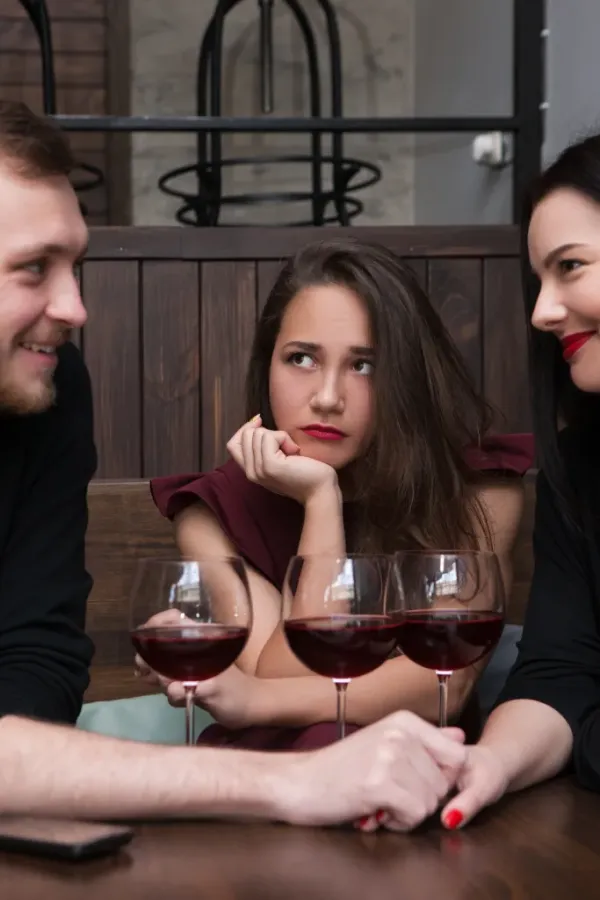 A woman in a red dress looking jealous while a man flirts with another woman at a table with three glasses of red wine.