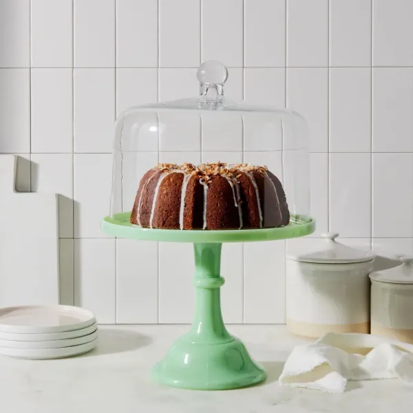 Bundt cake with white icing and toasted coconut on a green cake stand with a glass cloche.