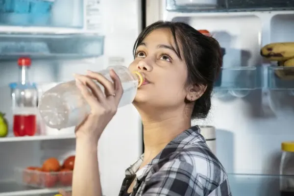 Minum air putih di tengah malam Young woman standing in front of open refrigerator and drinking cold water while suffering from a hot summer heat at home