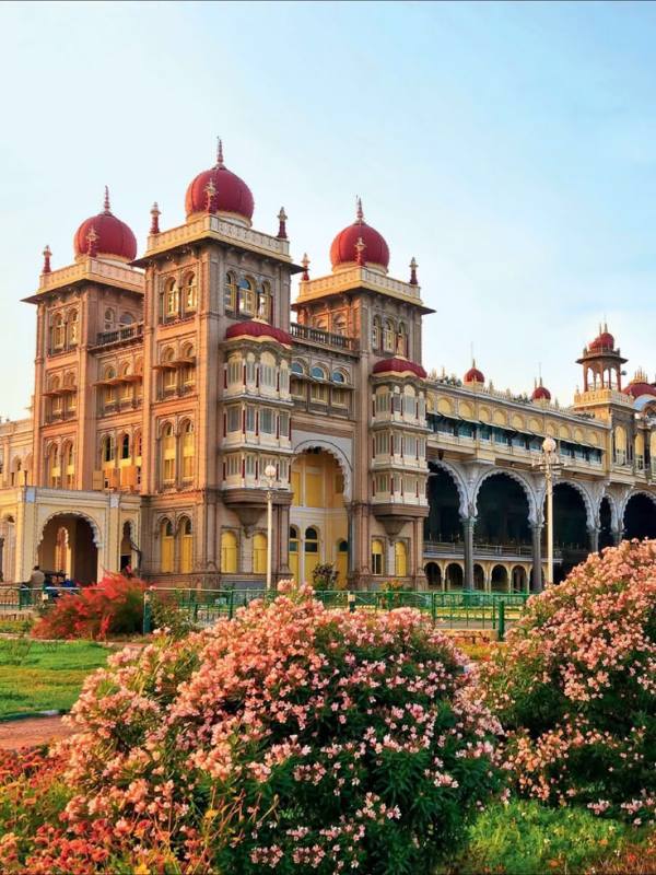 This may contain: an ornate building with red domes on top and pink flowers in the foreground, surrounded by greenery