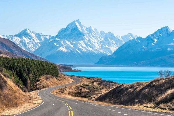 This may contain: an empty road with mountains in the background and blue water on one side, surrounded by grass and trees