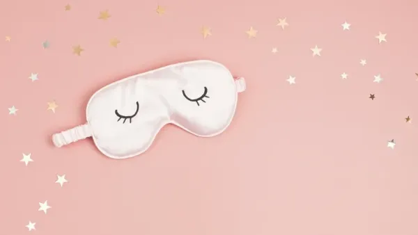 White sleep mask with closed eye design on a pink background with scattered gold stars.