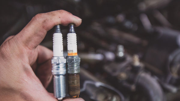 A man holding a new and old spark plug side-by-side, out of focus engine in the background