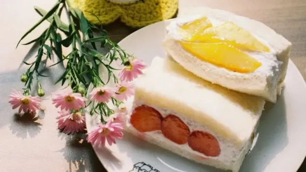 Soft white bread fruit sandwich filled with cream, strawberries, and mango slices, served on a plate with pink flowers on the side.