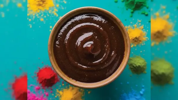 Bowl of thick brown chutney or sauce with colorful spice powders scattered around on a vibrant background.