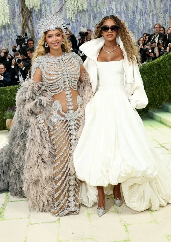 Beyoncé in a sparkling silver skeleton-like gown and feathery cape, standing next to Blue Ivy Carter in a white gown and jacket, at the 2026 Met Gala.