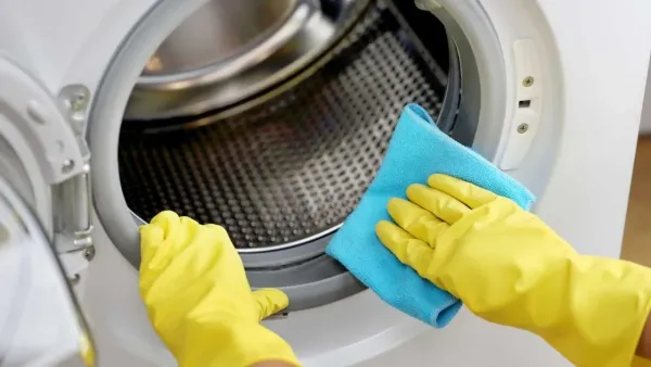 Close-up view of a hand wearing yellow gloves cleaning the drum of a washing machine with a blue microfiber cloth.