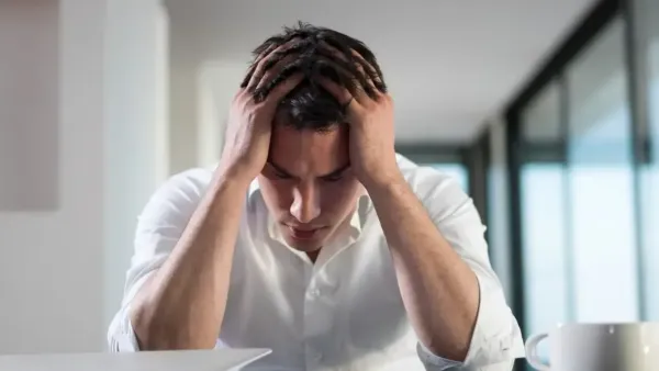 A man in a white shirt sits with his head in his hands, looking down in stress or frustration, with a laptop and a cup placed on the table in front of him.