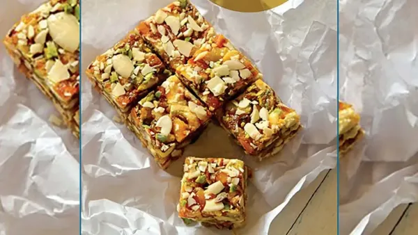 Square pieces of healthy dry fruit barfi or energy bars loaded with chopped almonds and pistachios on white parchment paper.
