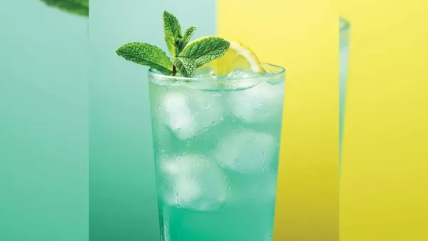 A tall glass filled with an icy teal mint mojito, garnished with a lemon slice and a fresh mint sprig on a green and yellow background.