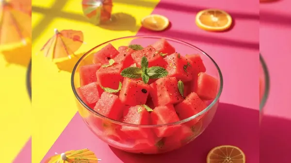 Juicy red cubed watermelon pieces sprinkled with black pepper and mint leaves in a glass bowl, decorated with mini paper umbrellas on a pink and yellow shadow background.