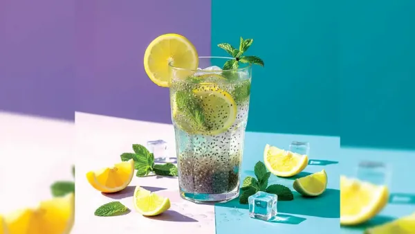 A tall clear glass of refreshing lime water filled with hydrated sabja seeds, sliced lemons, and mint leaves, placed on a pastel purple and blue background with ice cubes around it.