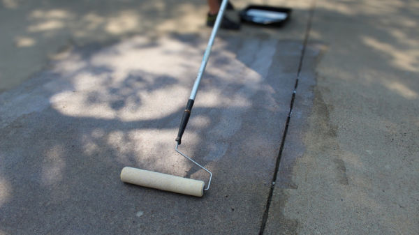 A person using a long pole and paint roller to apply driveway sealer