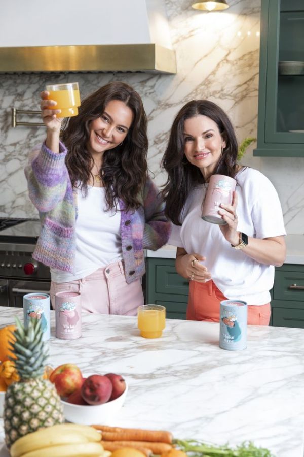 Emma Heming in a kitchen raising a glass with another individual fruit and beverage cans on the counter Credit: Make Time Wellness