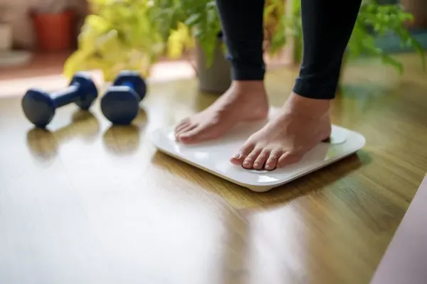 A close-up view of a young woman standing on a weighing scale, focusing on her feet and the display as she checks her weight. The scene highlights the importance of health and self-awareness in maintaining a balanced lifestyle.