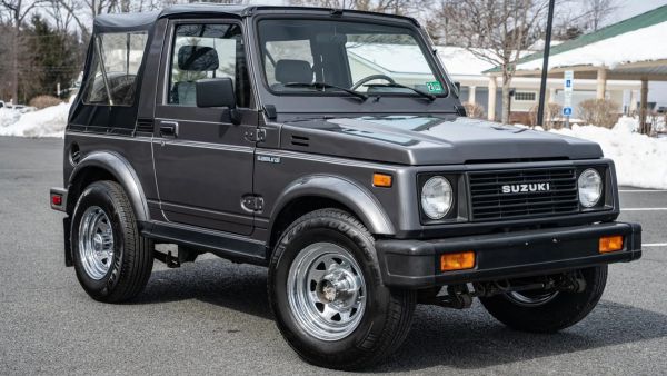 Gray Suzuki Samurai SUV with soft top parked on asphalt with snow in the background.
