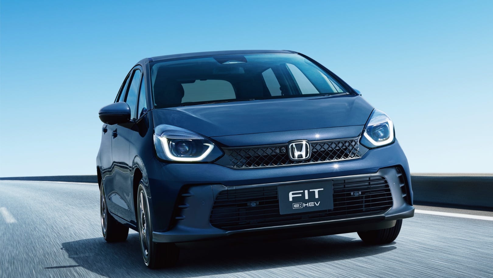 Blue Honda Fit HEV driving on a highway under clear sky.
