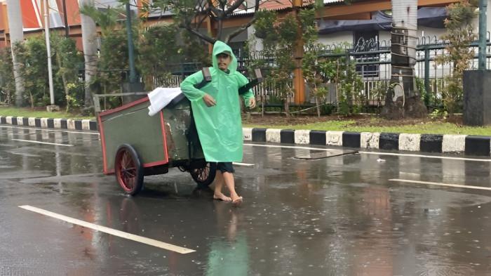 KEHUJANAN- Seorang pria yang tengah membawa gerobak tampak kehujanan dan harus menggunakan jas hujan saat kegiatan di luar rumah. Ia terlihat tidak menggunakan alas kaki saat melintasi jalan yang basah.