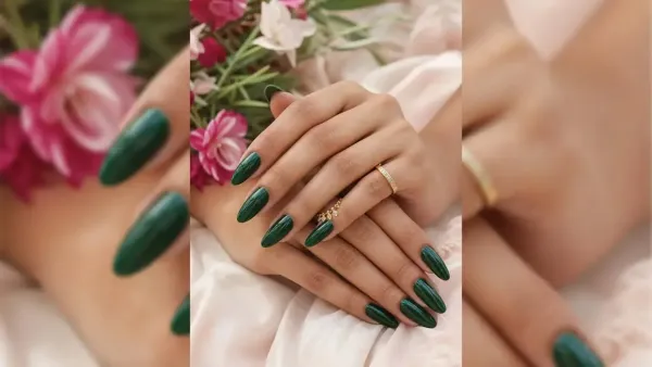 Woman displaying long glossy dark green manicured nails with gold ring accents placed against a soft floral background.