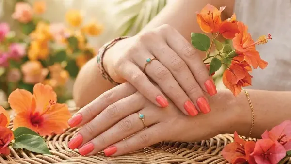 Woman displaying glossy bright coral peach manicured nails with turquoise rings surrounded by vibrant orange tropical flowers.
