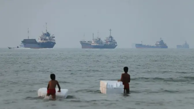 Dua anak laki-laki bermain di perairan dangkal di lepas pantai Bandar Abbas, Iran, dengan latar kapal-kapal yang melintas di Selat Hormuz, Selasa (30/6/2026). (Dok. Amirhosein Khorgooi/ISNA via AP)
