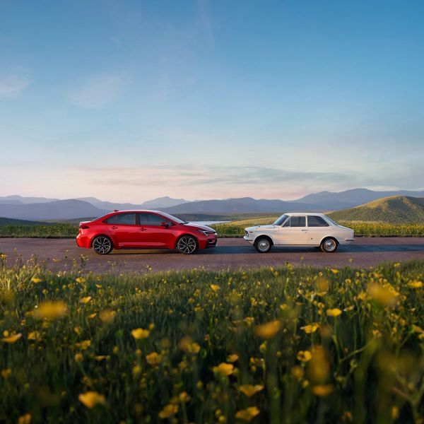 Red modern sedan and white vintage compact car parked facing each other on a rural road with mountains in the background.