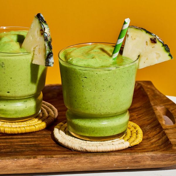 Two glasses filled with pineapple green smoothie, garnished with a pineapple wedge and a green and white striped straw Credit: Photographer: Morgan Hunt Glaze, Prop Stylist: Phoebe Hausser, Food Stylist: Emily Nabors Hall