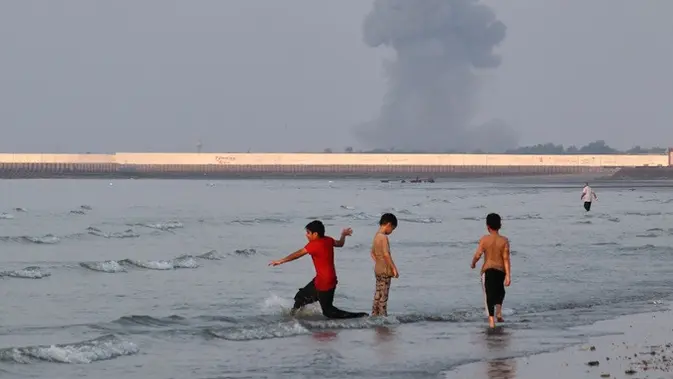 Tiga anak laki-laki bermain di perairan dangkal Selat Hormuz saat kepulan asap akibat ledakan terlihat di lepas pantai Bandar Abbas, Iran, Senin (13/7/2026). (Dok. Razieh Poudat/ISNA via AP)