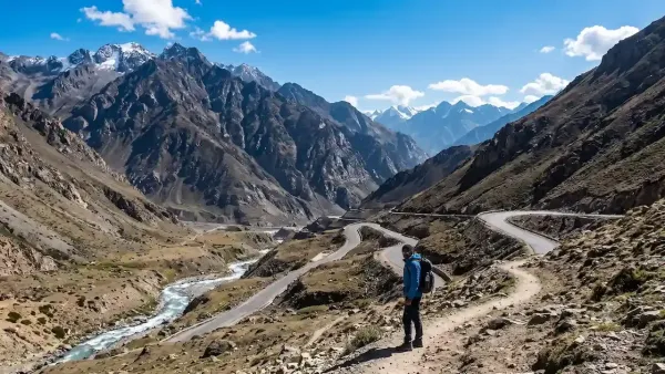 Male trekker with backpack standing on mountain trail overlooking winding valley highway and flowing river in Himalayas.