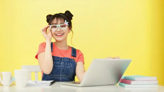 Siswa perempuan Asia muda yang tersenyum dengan kacamata berwarna cerah dan duduk di meja dengan laptop (Foto dok : Freepik/pressfoto).